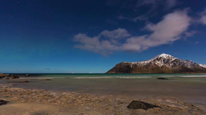 Beach with a sandy and rocky foreground calm sea and a coastal mountain under a partially cloudy night sky