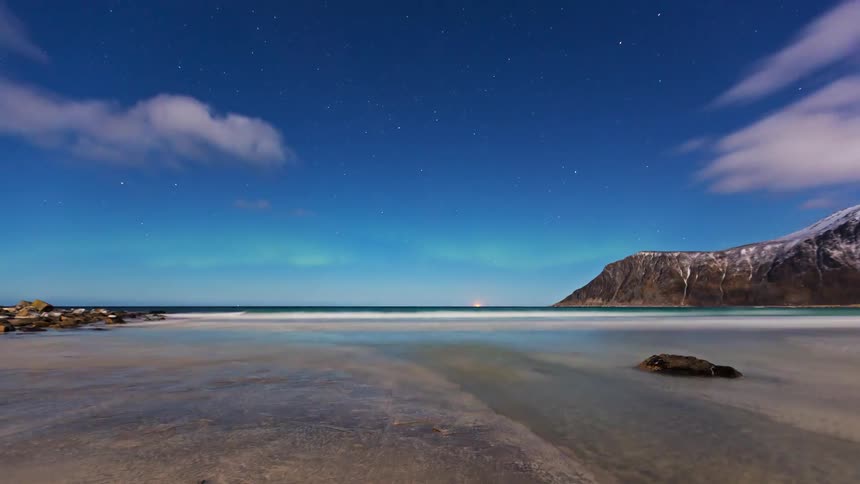 Mountains on the coast and the aurora borealis glows green in the sky as clouds move overhead