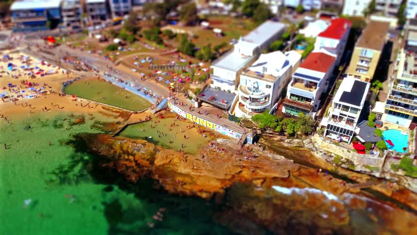 This high angle tilt shift view shows people swimming in an ocean pool and relaxing on the sand at beach next to a row of white coastal apartments and rugged rocks