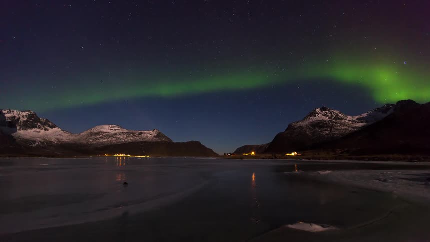 Green aurora borealis lights shimmer across a starry night sky over snow covered mountains and a frozen lake with distant village lights reflecting on the icy surface