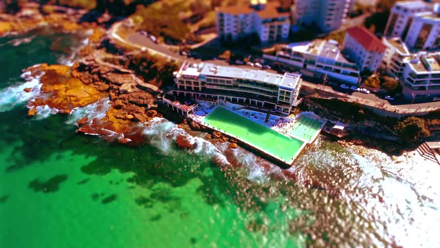 High angle drone shot captures the iconic bondi icebergs pool nestled against rugged rocks as bright turquoise ocean waves crash against the concrete walls while people swim and sunbathe near the white coastal buildings and winding road