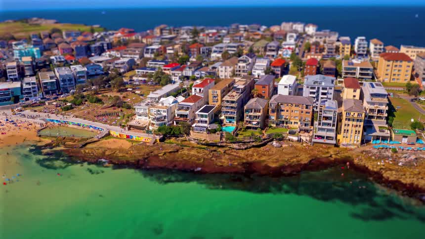 An aerial view captures a vibrant coastal neighborhood perched along rocky shores where clear turquoise water meets a dense cluster of seaside buildings