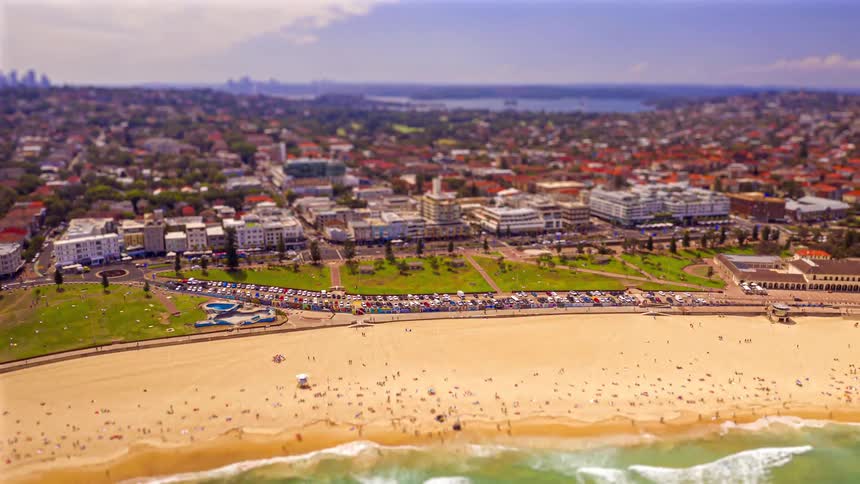 A high angle view shows a crowded sandy beach with white waves rolling in from a green ocean as a coastal city stretches into the distance under a hazy sky