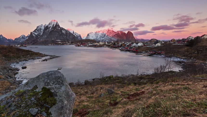 A quiet coastal village sits beside a calm inlet framed by snowcapped mountains glowing pink in the soft light of sunset