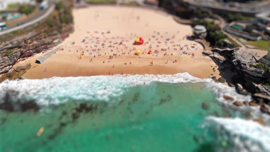 A miniature style aerial view shows waves crashing onto a crowded beach with vibrant turquoise water