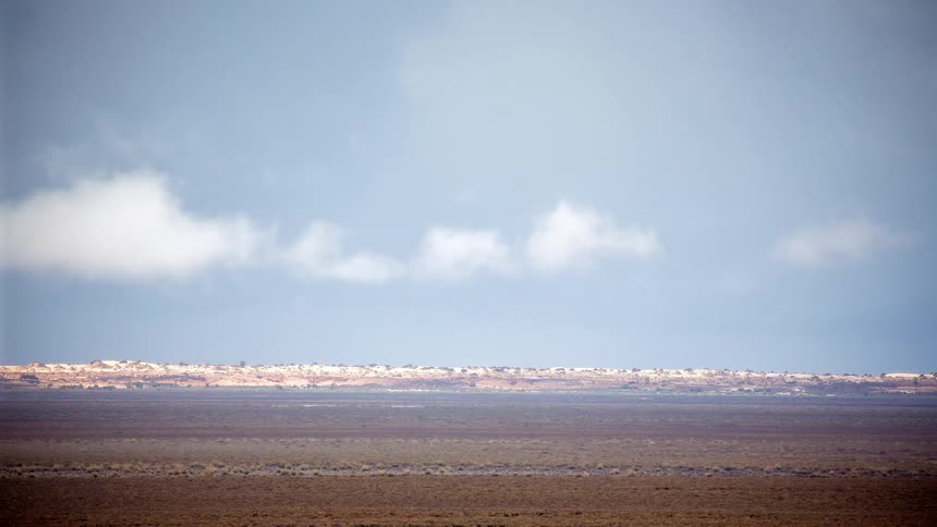 This timelapse video captures soft white clouds drifting across a pale blue sky below the horizon a vast and desolate desert landscape stretches out in muted brown and tan tones
