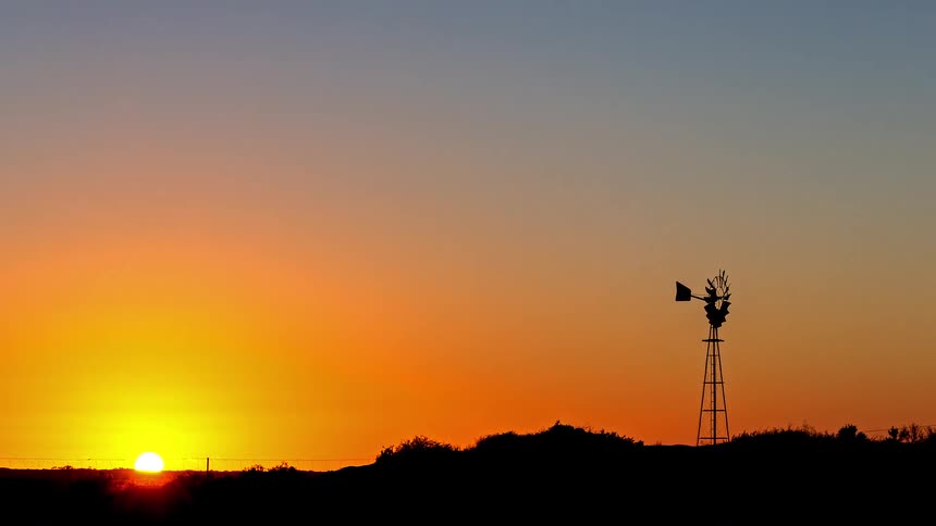 Sun rising on the horizon with a windmill silhouette over elevated terrain