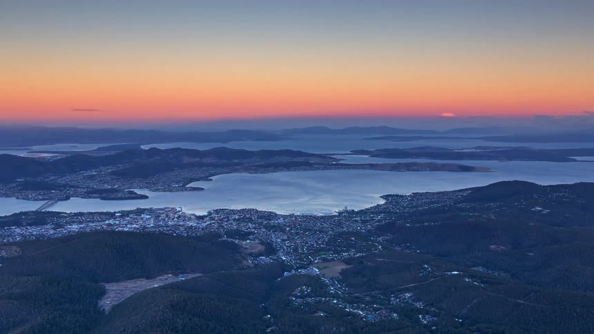 A sweeping aerial view looks across rolling hills to a broad lake surrounded by towns bathed in soft early morning light under a clear sky