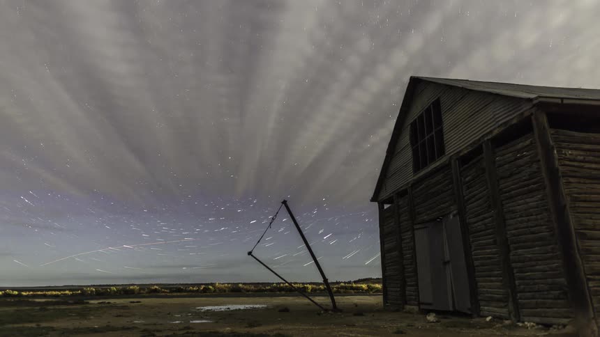 Star trails in the night sky over an open field with a wooden barn on the right