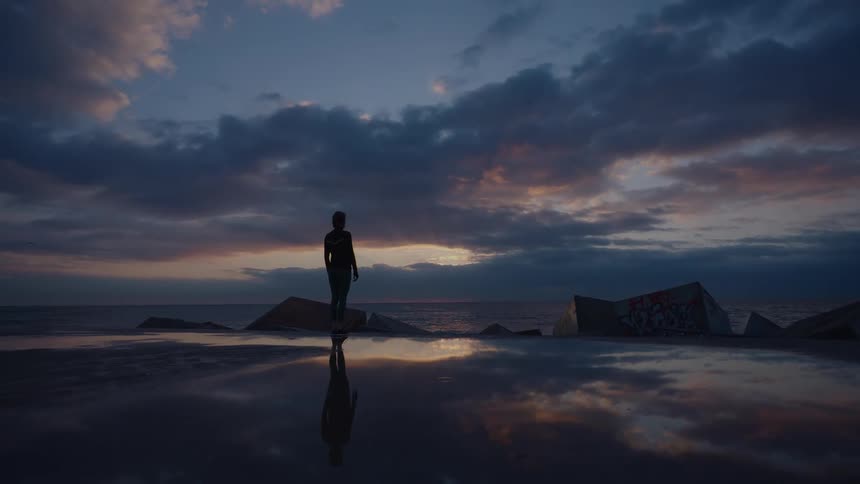 A lone figure stands silhouetted on a reflective shoreline at dusk gazing toward the ocean beside angular rocks beneath dramatic darkening clouds