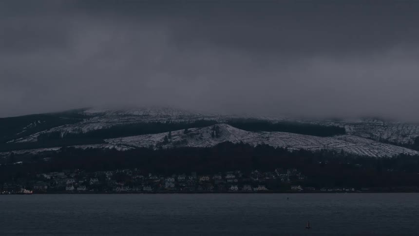 Coastal mountain partially covered with snow with an urban area along the waterfront under an overcast sky