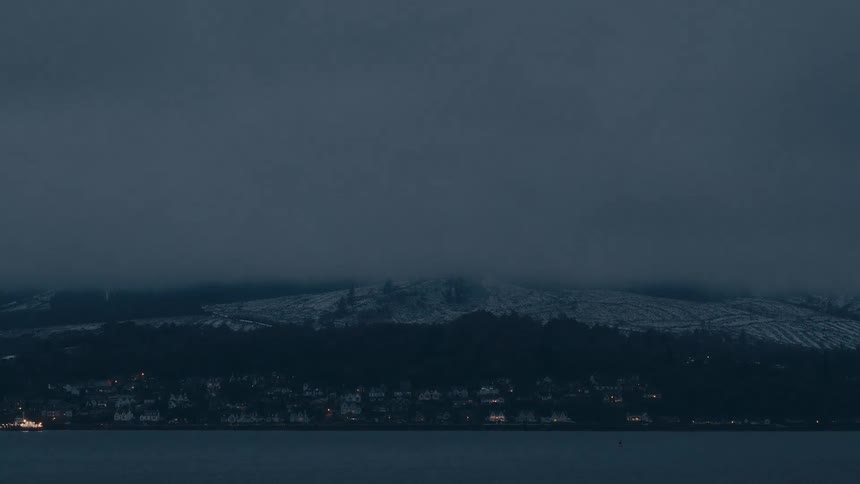 Timelapse of coastal mountain under dense clouds with urban lights turning on along the waterfront at nightfall