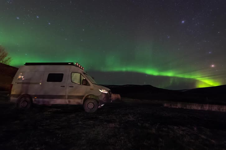 Van parked along the coast as the green lights of the aurora borealis glow over mountains in a starry sky