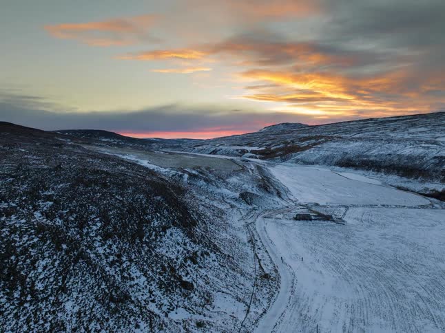 An aerial view shows a waterfall cascading down a dark rocky cliff into a snowy valley during a sunset with vibrant orange and blue clouds in the sky