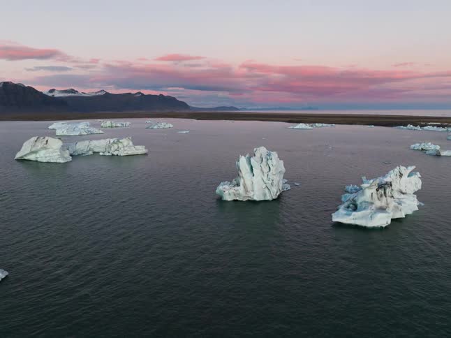 An aerial shot glides over a vast glacial lagoon revealing jagged white and turquoise icebergs floating in dark tranquil waterthe scene is set against a backdrop of snowdusted mountains and a soft twilight sky streaked with pink and orange clouds