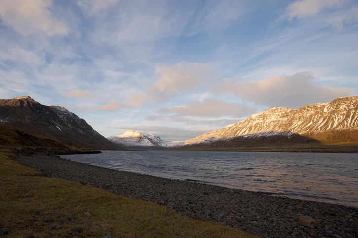 Clouds move through the sky above a body of water bordered by a rocky shore and snow dusted mountains