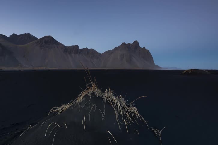A wide shot of a black sand beach at dawn with a small dune of yellowed grass in the foreground in the background a massive jagged mountain range sits under a clear dim sky as the camera slowly pulls back