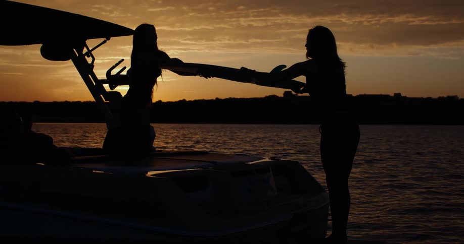 Two women are seen in silhouette on a boat at sunset with one woman handing a surfboard to the otheras the sun sets over the water the golden sky creates a beautiful backdrop for the scene of friends enjoying an evening of surfing