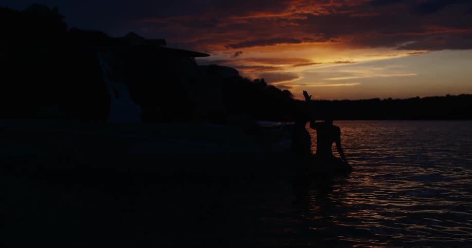 A silhouette of a person raising their hand and waving toward the horizon as the sun dips below the clouds the second line focuses on the calm water in the foreground which reflects the deep orange and purple hues of the evening sky