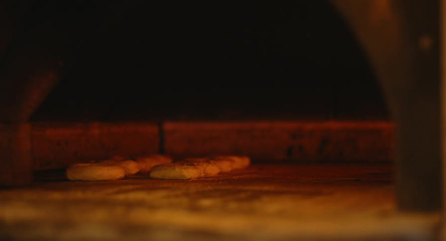 A baker places fresh dough on the stone floor inside a wood fired oven in a dimly lit bakery kitchen