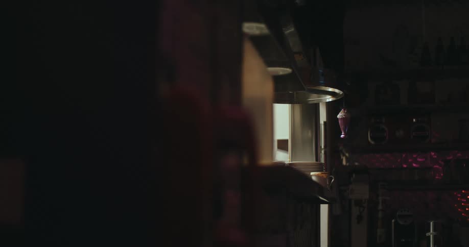 A view from a dark corner shows stacked white plates on a metal shelf in a dimly lit kitchen with a small pink object hanging nearby