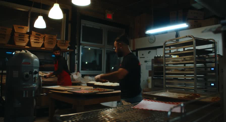 Two bakery staff working at their stations inside a dimly lit commercial bakery