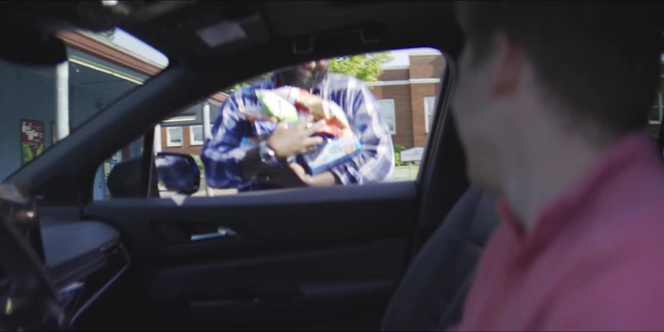 A view from inside a car shows a man in a plaid shirt walking up to the passenger side window carrying several bags of snacks and groceries