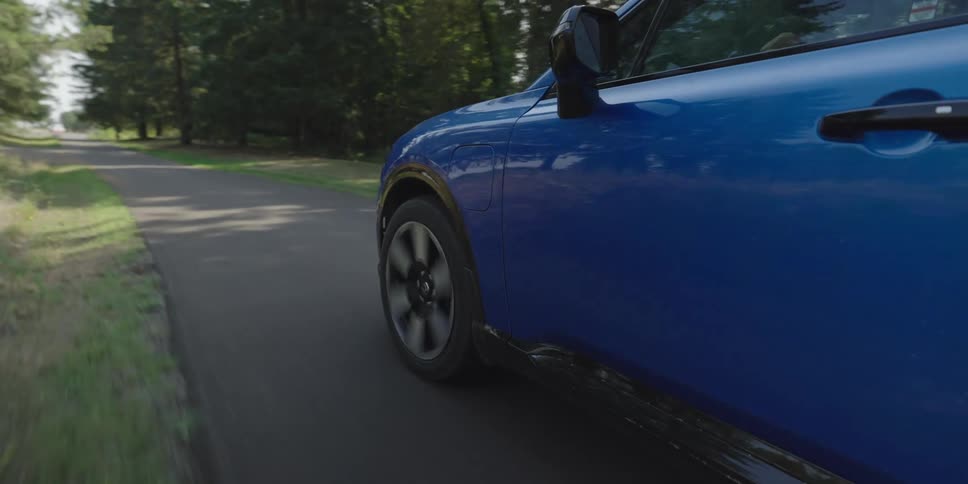 A blue car drives quickly along a narrow asphalt road winding through a dense forest of green pine trees with its front wheel spinning and reflecting the bright daylight on its metallic surface