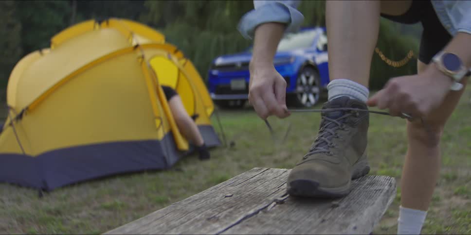 A close up view shows a person placing their foot on a wooden bench to tie the laces of a brown hiking boot at a campsite while another person organizes gear near a yellow tent and a blue suv in the background