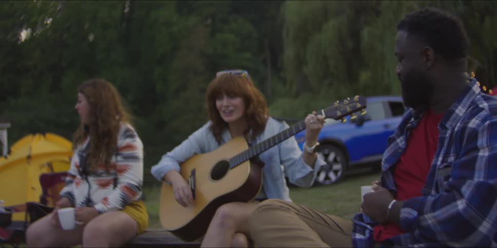 Young adult woman singing and playing acoustic guitar sitting on a wooden bench with people around her drinking from ceramic cups with tents and cars on the background outdoors in a natural area during dusk or dawn