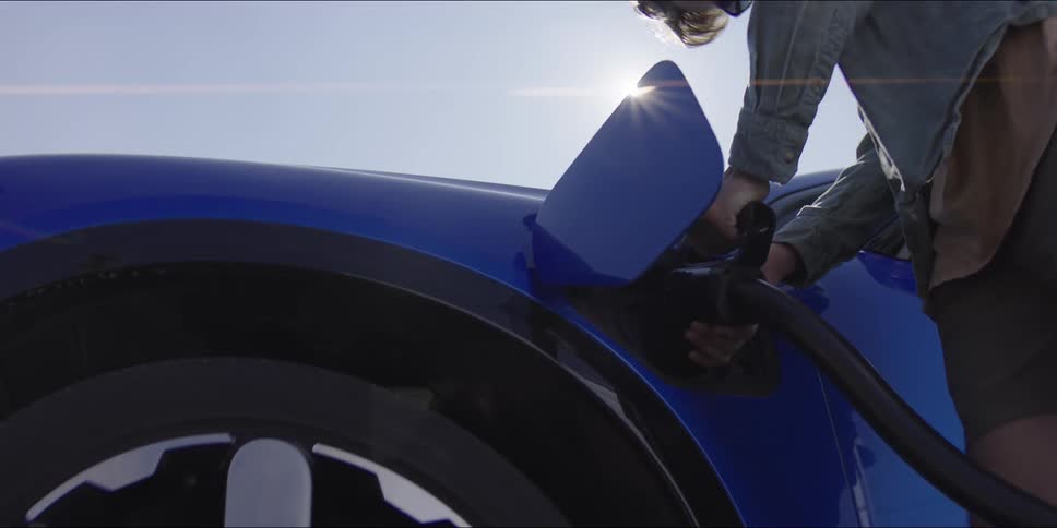 A person wearing a denim shirt reaches toward a blue electric car and plugs a charging cable into the side port under a clear sky
