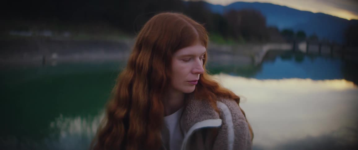 A close up shows a woman with long wavy red hair wearing a beige fleece jacket as she looks out over a calm body of water with mountains in the background during twilight
