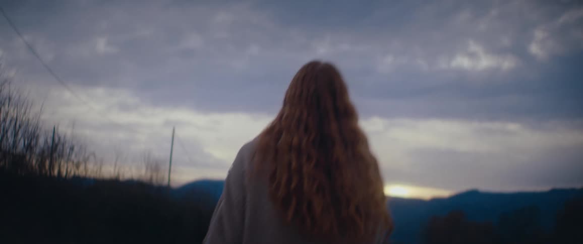 A woman with long hair walking alone outdoors under a cloudy evening sky