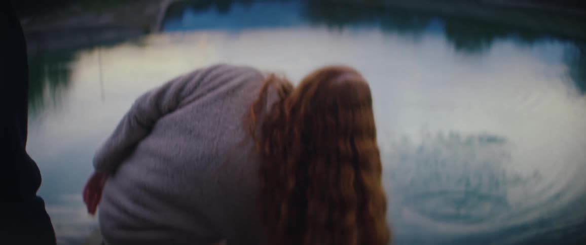 A young woman with long red hair skips a stone across the water and turns back with a bright smile