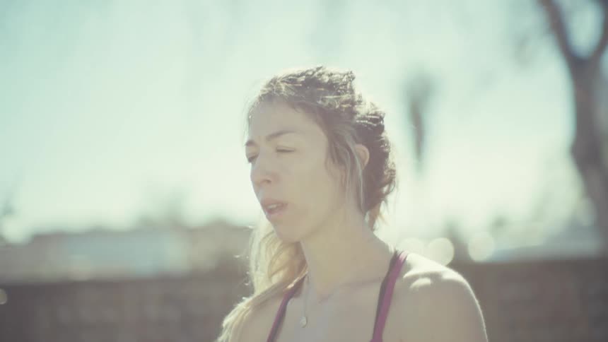A pregnant woman in athletic wear stands in a sunlit outdoor setting gently cradling her belly while practicing mindfulness