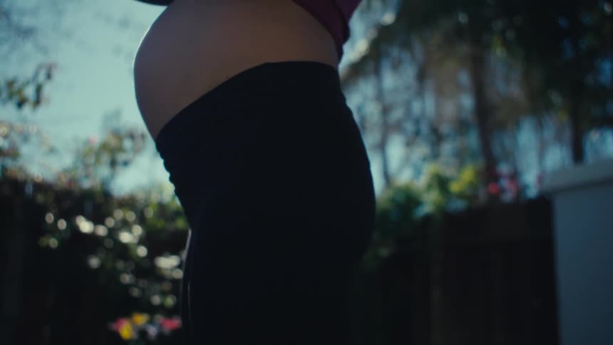 Adult pregnant woman on a lawn squatting while holding a kettle bell outdoors during day time
