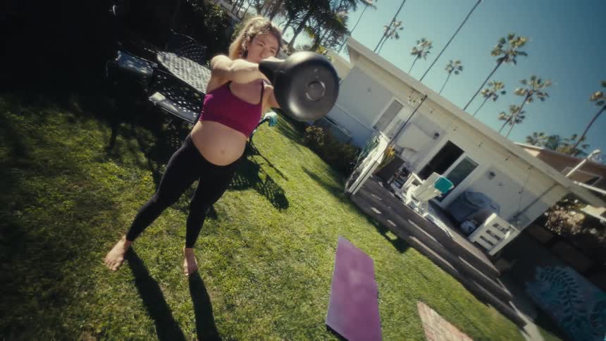 Expectant mother swings a weight while exercising barefoot in her backyard on a summer day