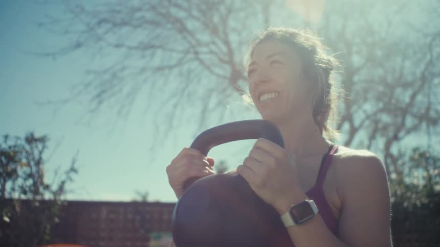 A young woman performs kettlebell swings during an outdoor workout on a sunny day and she wears a purple tanktop and a smart watch while exercising in a backyard with trees and a clear blue sky in the background