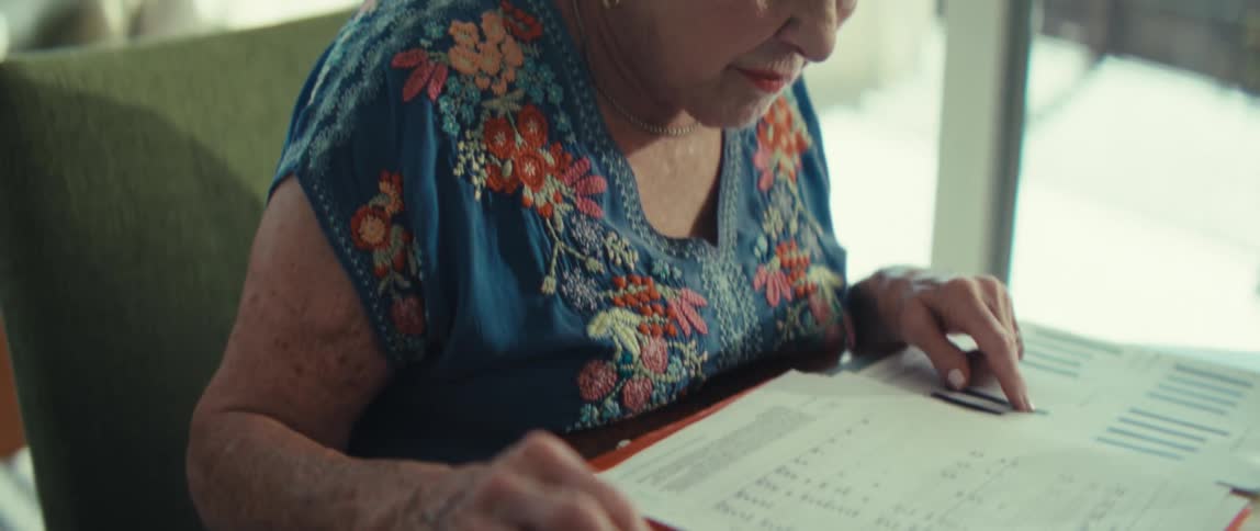 A senior woman uses a white and orange desktop calculator to process financial information from open documents on a wooden table