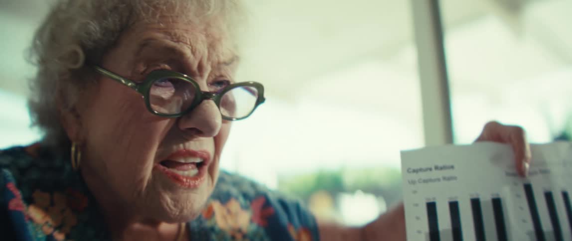 A close up shows an older woman wearing glasses and a floral shirt as she holds and points to a document featuring financial bar charts labeled capture ratios