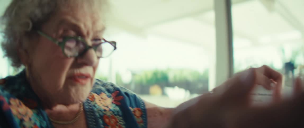 An older woman with curly white hair wearing glasses and a blue floral shirt holds a document with charts while talking with a serious expression inside a bright room