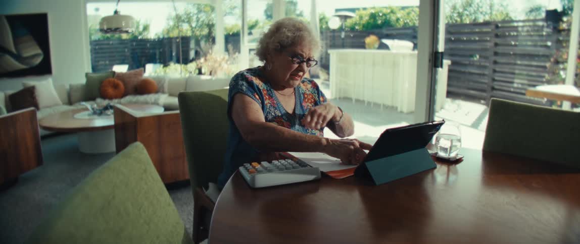 Elderly woman sitting in front of a table with a calculator and a tablet computer with an angry and nervous facial expression indoors during day time