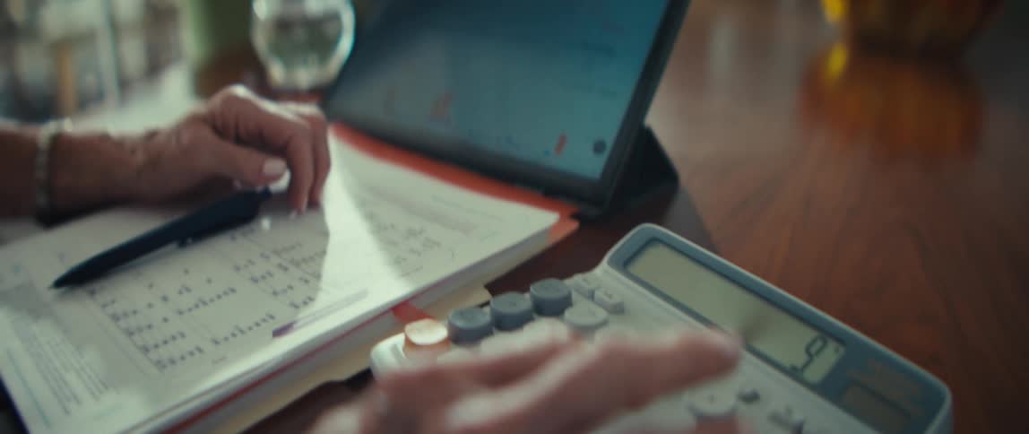 A close view shows the hands of an older woman as she uses a calculator next to a tablet displaying financial data on a wooden table