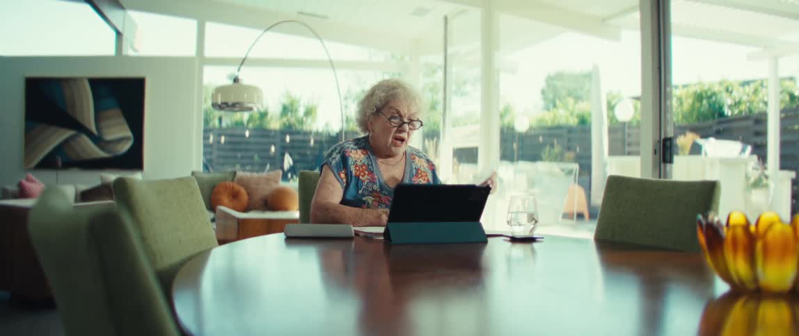 An elderly woman sits at a round wooden table in a bright modern house while holding up a document with charts to a tablet camera during a video call