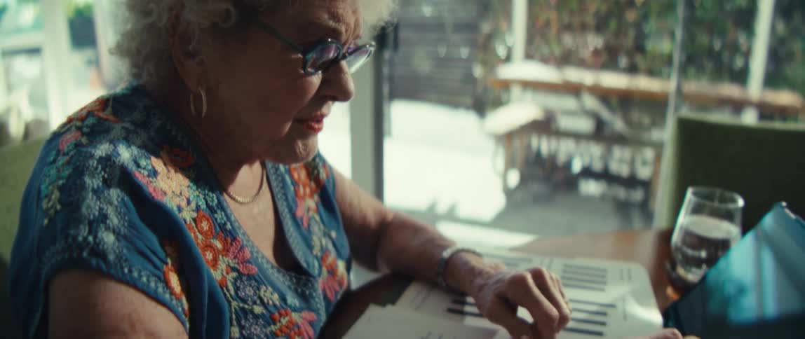 An elderly woman wearing glasses sitting at a desk and using a tablet pc in the living room at midday