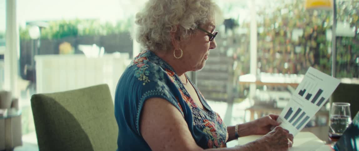 An elderly woman sits at a dining table while reviewing a document