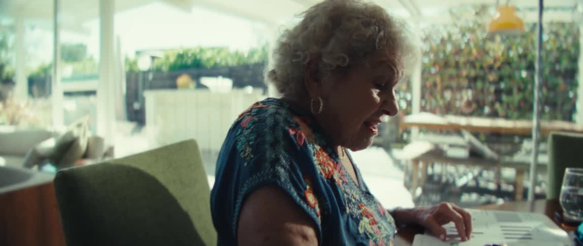 An elderly woman sits at a wooden dining table in a sunlit modern house while she carefully writes on paper documents near a tablet