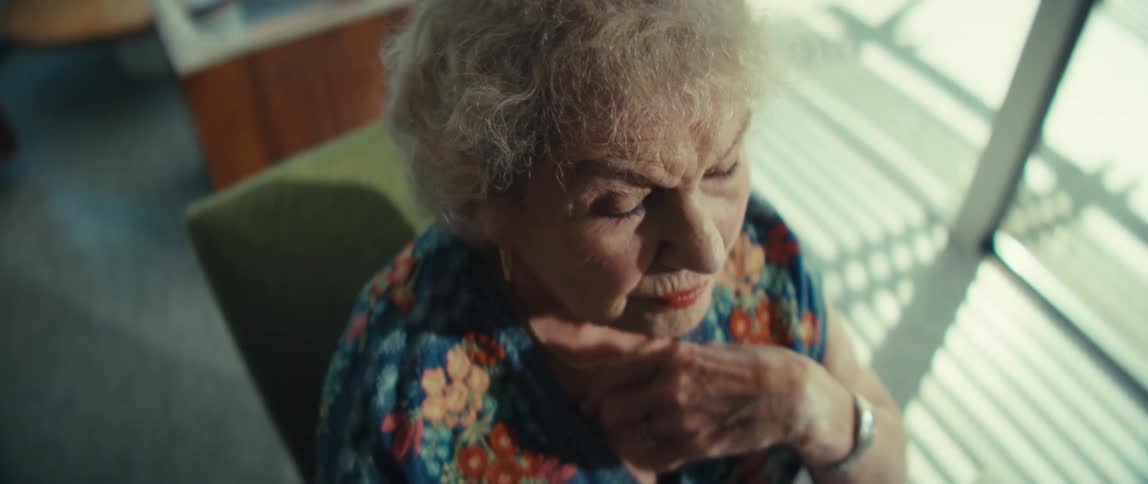An elderly woman sits at a wooden table looking intensely at financial documents with charts while holding a pen