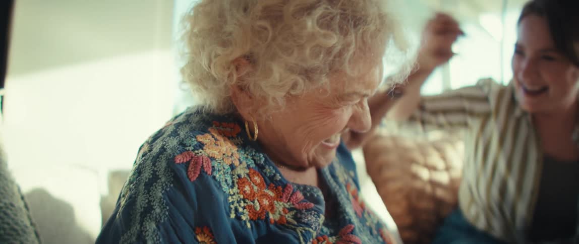 An elderly woman and a young woman sitting on a couch indoors at midday laughing and highfiving while enjoying a conversation