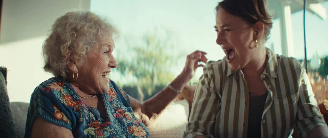 A grandmother and granddaughter laugh while playing a handheld video game together in a modern living room